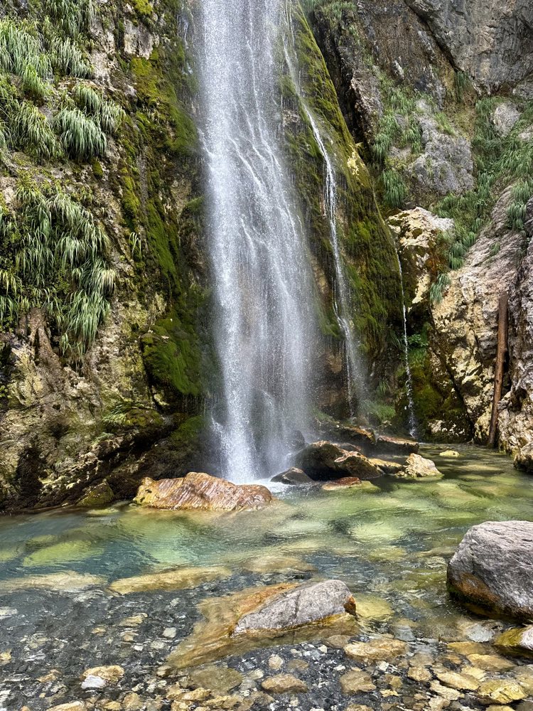A waterfall pouring off a mossy cliff into a turquoise pool in Theth